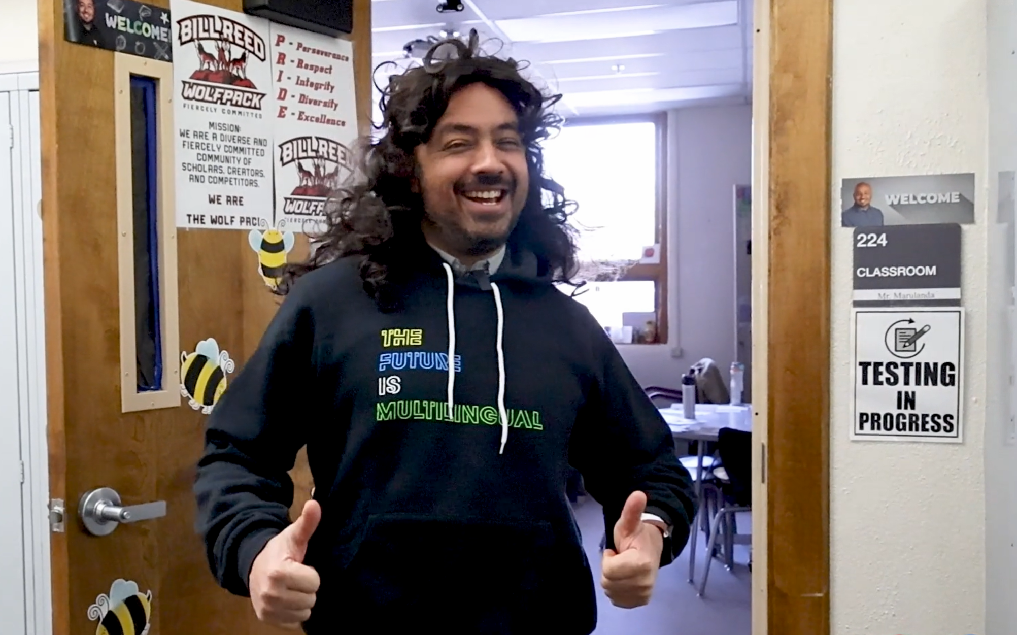 A teacher wearing a long, dark curly wig gives two thumbs up. 